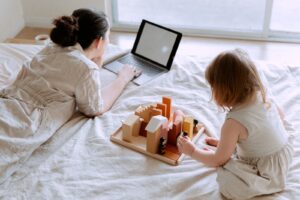 From above of young female in casual clothes lying on bed and using laptop while daughter sitting on bed and playing with wooden blocks and toys on sunny morning